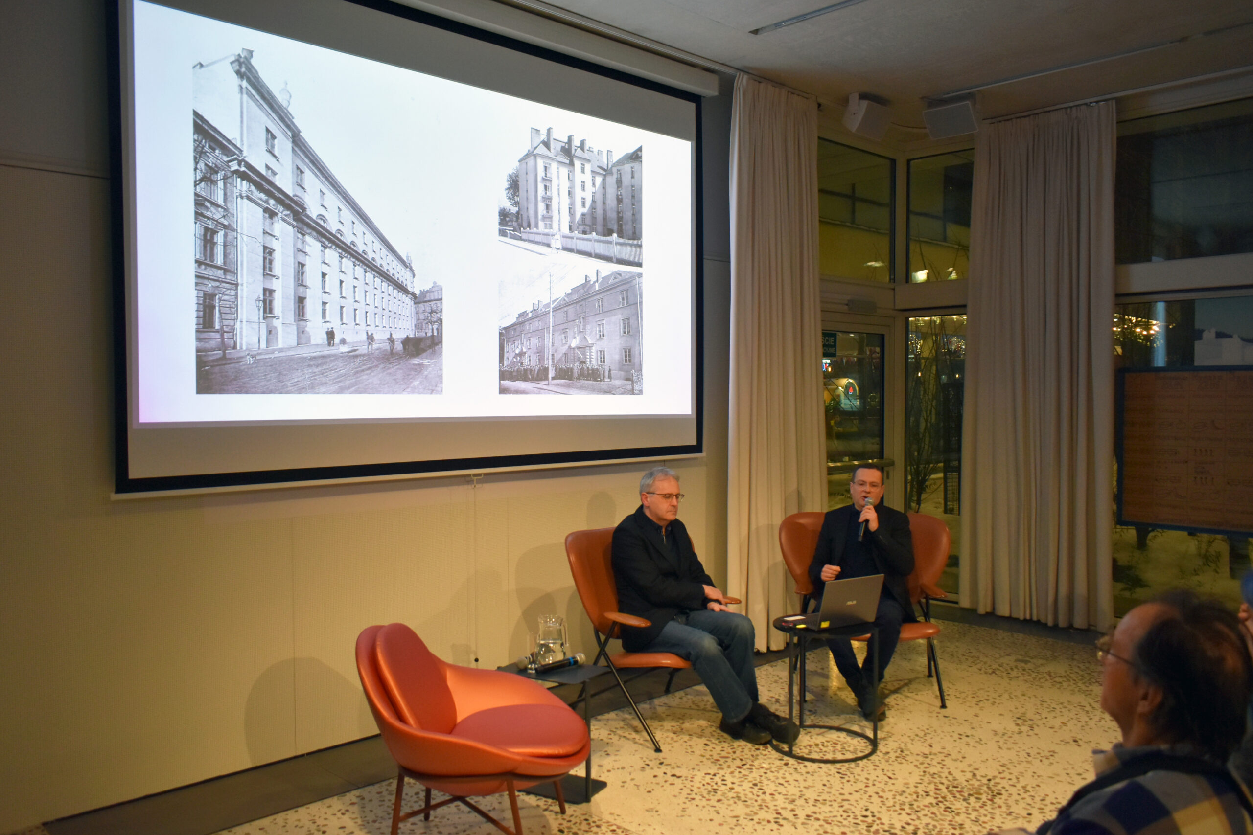 Dwóch mężczyzn siedzi na pomarańczowych fotelach przed dużym ekranem projekcyjnym w nowoczesnej sali. Na ekranie wyświetlane są trzy czarno-białe fotografie historycznych kamienic. Jeden z mężczyzn mówi do mikrofonu, trzymając przed sobą laptopa.